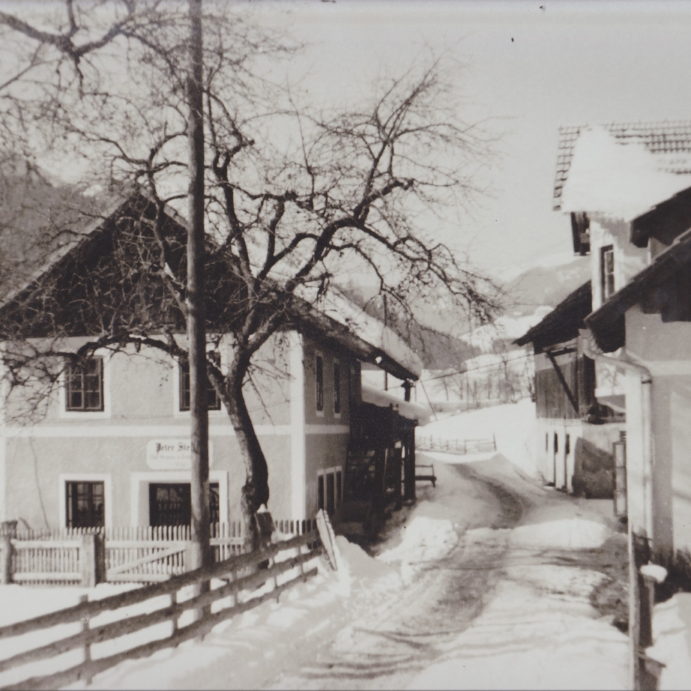 Schmiedewerkstatt und Wohnhaus von 1933 Ganz links im Bild befindet sich das Kraftwerkhaus. Rechts daneben das große Werkstattgebäude mit dem giebelseitigen Eingang, darüber die Haus- und Berufsbezeichnung “Peter Steiner”, “Huf-Wagenschmiede” und die Hausnummer 12. Straßenseitig ist der Stiegenaufgang in das Obergeschoss noch erhalten. Gegenüber der Straße zeigt die Fotografie nach 1933 das umgebaute Wohnhaus mit dem dahinterliegenden Wirtschaftsgebäude.